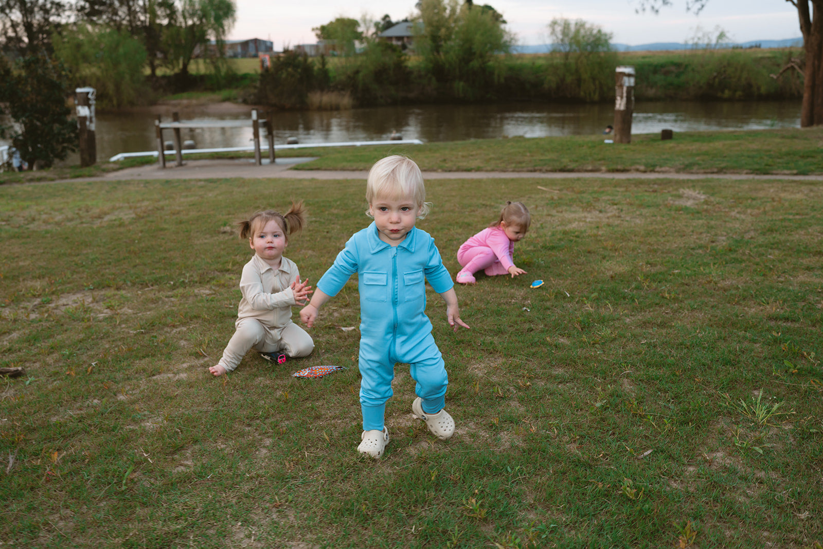 Three children playing on grass near a pond with trees and a house in the background.