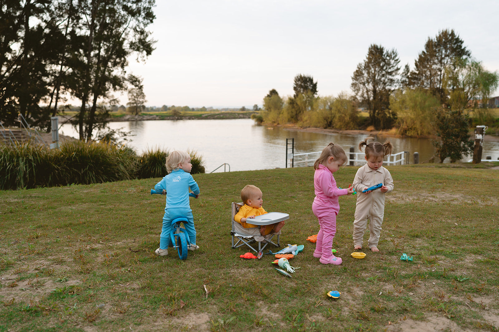 Four children playing on a grassy area near a lake with trees in the background.