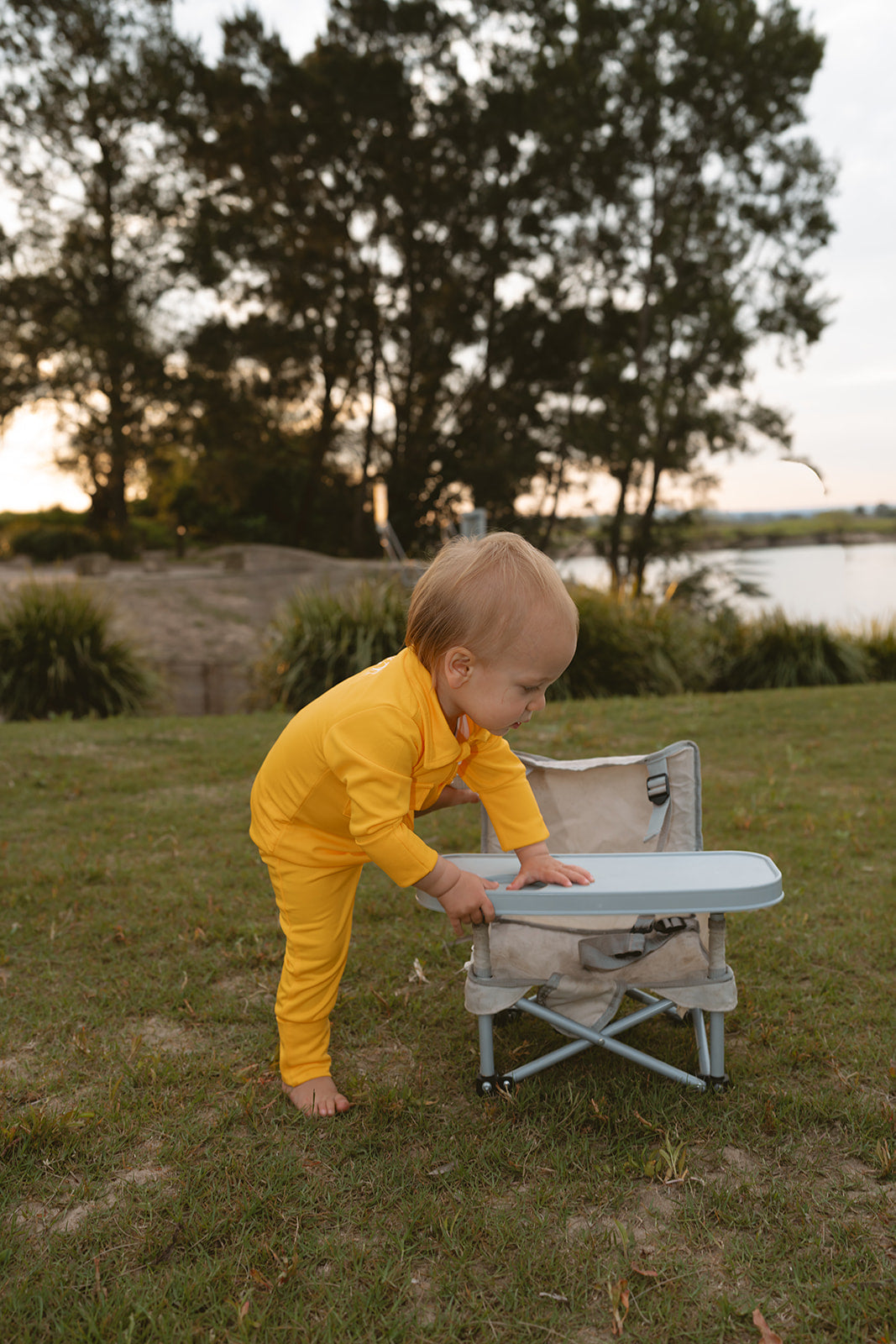 Child in yellow outfit interacting with a small table outdoors near trees and water.