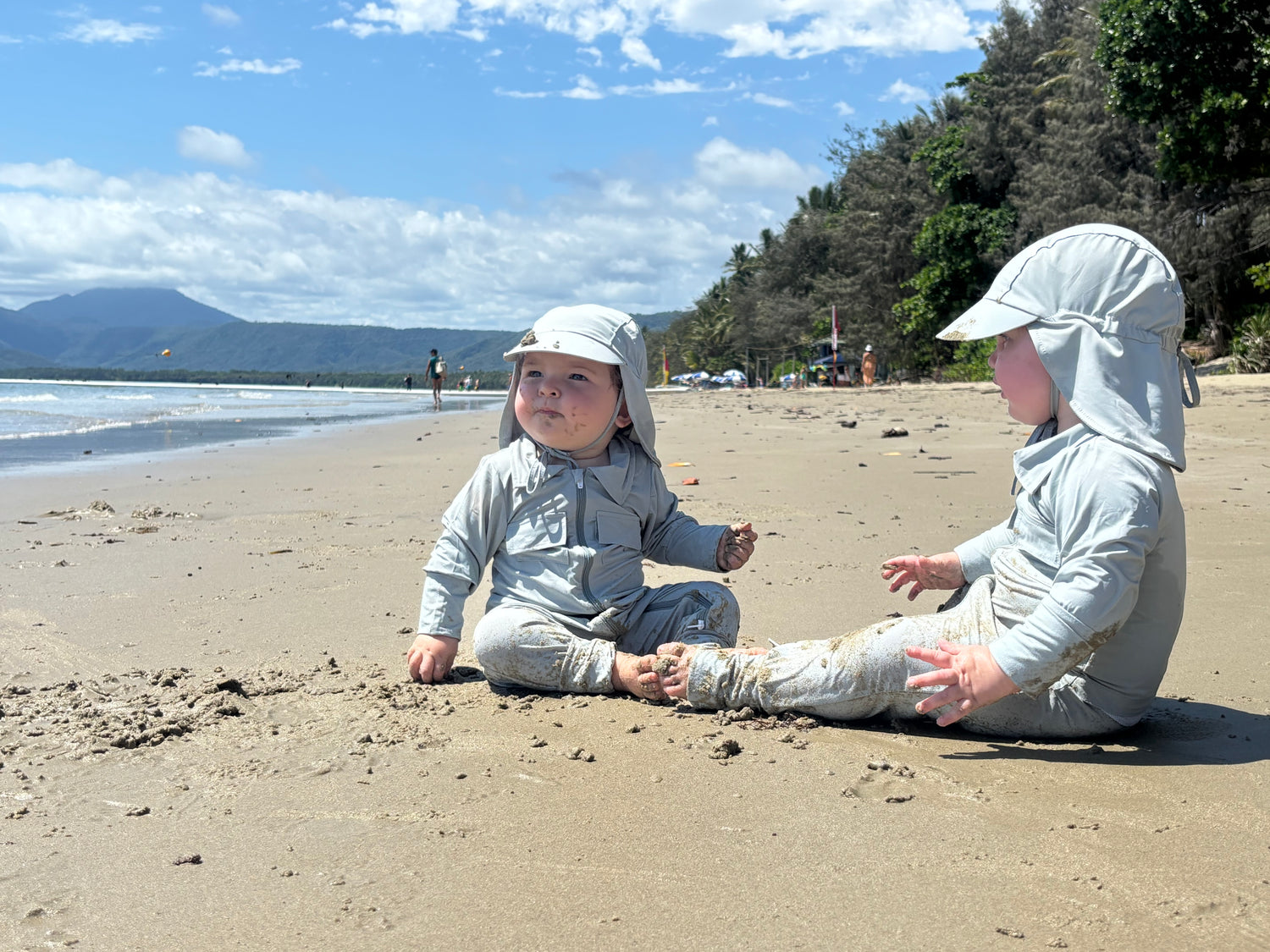 Two children in light clothing sitting on a sandy beach with mountains and trees in the background.